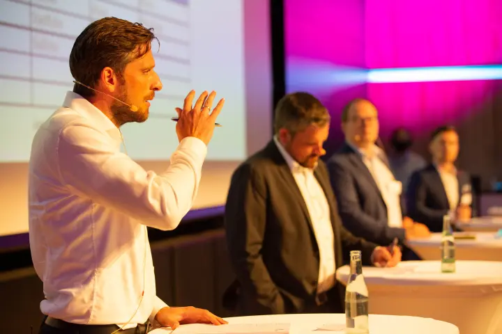 Mann mit Headset-Mikrofon, im weißen Hemd, steht links im Vordergrund auf einer Bühne, gestikuliert mit einem Stift in der Hand und hält eine Präsentation vor einem Publikum, das im Halbdunkeln an runden, weiß gedeckten Tischen sitzt. Die Bühne ist im Hintergrund pink und violett beleuchtet.