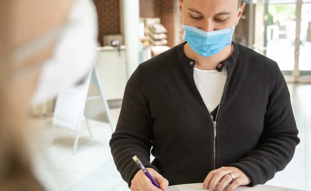 Mann mit Gesichtsmaske unterschreibt Dokument an einem Tisch.