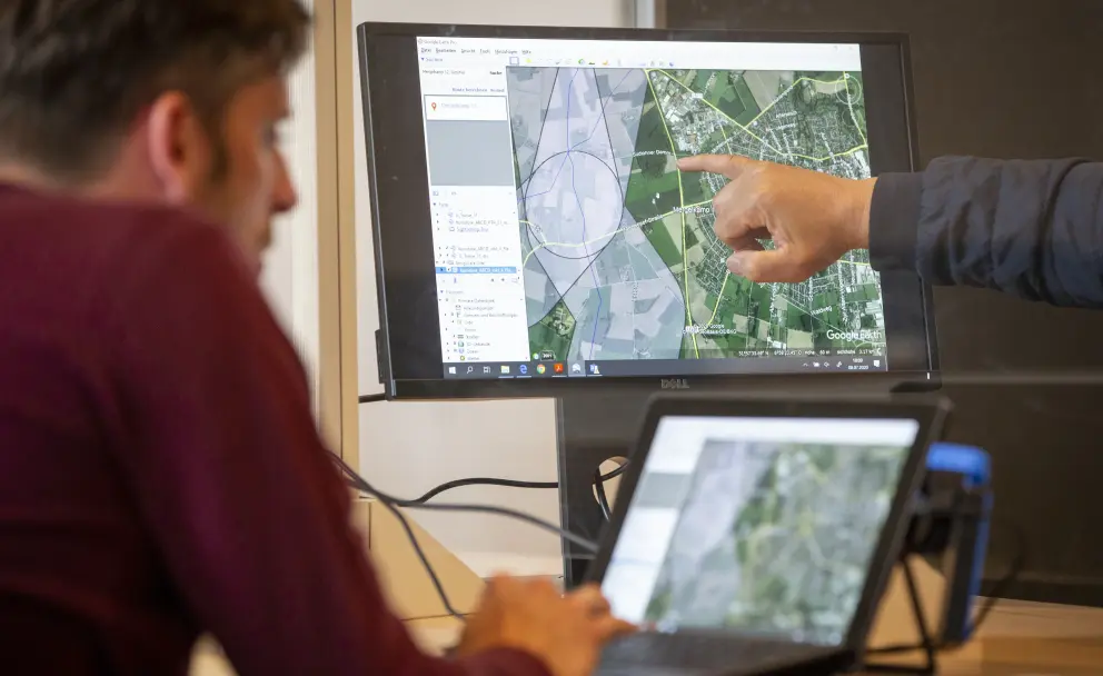 Eine Hand zeigt auf eine Luftaufnahme mit eingezeichneten Routen am Bildschirm, während eine zweite Person am Laptop tippt.