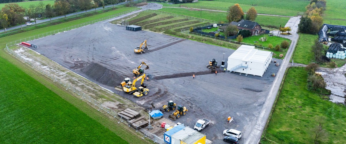 Das Bild zeigt eine Luftaufnahme einer aktiven Baustelle in einer ländlichen Umgebung, umgeben von grünen Feldern und Wiesen.
Beschreibung des Bildes
Das zentrale Element ist eine große, rechteckige, grau-braune Schotterfläche. Auf dieser Baustellenfläche befinden sich mehrere Baumaschinen, darunter große gelbe Bagger, sowie kleinere Fahrzeuge wie Lieferwagen und PKWs. Im unteren Bereich der Fläche steht ein blauer Container und eine weiße Zeltstruktur im mittleren rechten Bereich.
Die Baustelle ist klar abgegrenzt von der umliegenden Landschaft. Die Umgebung ist geprägt von weitläufigen grünen Wiesen und Feldern. Im oberen Teil des Bildes verläuft eine Straße, gesäumt von Bäumen, und weiter hinten sind einige Gebäude und Höfe erkennbar. Der Himmel ist bedeckt und grau.

