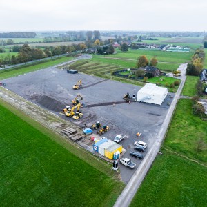 Das Bild zeigt eine Luftaufnahme einer aktiven Baustelle in einer ländlichen Umgebung, umgeben von grünen Feldern und Wiesen.
Beschreibung des Bildes
Das zentrale Element ist eine große, rechteckige, grau-braune Schotterfläche. Auf dieser Baustellenfläche befinden sich mehrere Baumaschinen, darunter große gelbe Bagger, sowie kleinere Fahrzeuge wie Lieferwagen und PKWs. Im unteren Bereich der Fläche steht ein blauer Container und eine weiße Zeltstruktur im mittleren rechten Bereich.
Die Baustelle ist klar abgegrenzt von der umliegenden Landschaft. Die Umgebung ist geprägt von weitläufigen grünen Wiesen und Feldern. Im oberen Teil des Bildes verläuft eine Straße, gesäumt von Bäumen, und weiter hinten sind einige Gebäude und Höfe erkennbar. Der Himmel ist bedeckt und grau.

