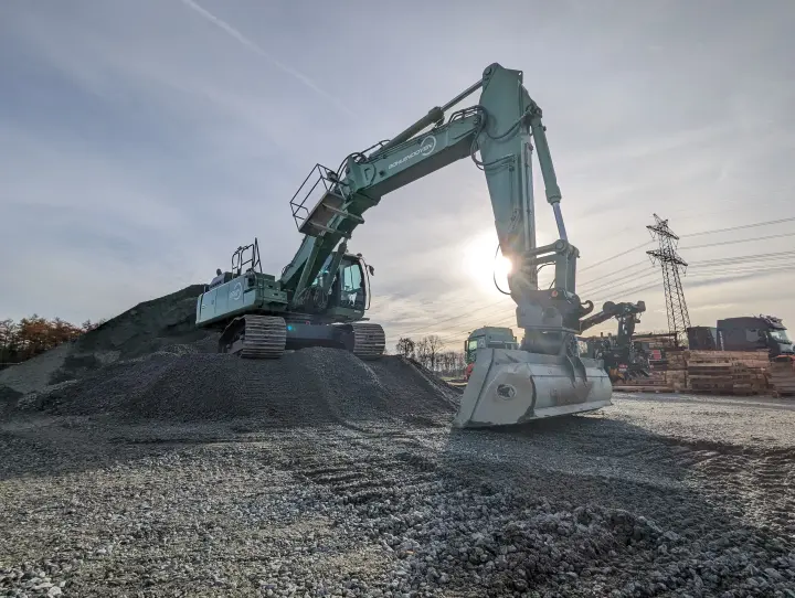 Grüner Raupenbagger mit Schaufel, auf einem Haufen Schotter, vor Freileitungsmasten bei Sonnenschein.