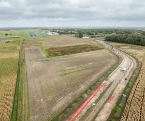 Luftaufnahme einer Großbaustelle für einen Erdkabelgraben, der durch landwirtschaftliche Felder verläuft, mit rotem Rohrmaterial und Baugeräten. Im Hintergrund sind die Landebahn und Gebäude eines kleinen Flugplatzes sichtbar, umgeben von Wald und Ackerland.