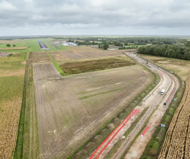 Luftaufnahme einer Großbaustelle für einen Erdkabelgraben, der durch landwirtschaftliche Felder verläuft, mit rotem Rohrmaterial und Baugeräten. Im Hintergrund sind die Landebahn und Gebäude eines kleinen Flugplatzes sichtbar, umgeben von Wald und Ackerland.