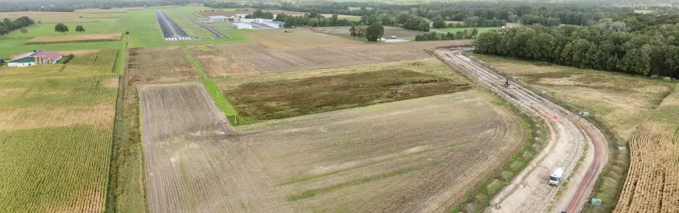 Luftaufnahme einer Großbaustelle für einen Erdkabelgraben, der durch landwirtschaftliche Felder verläuft, mit rotem Rohrmaterial und Baugeräten. Im Hintergrund sind die Landebahn und Gebäude eines kleinen Flugplatzes sichtbar, umgeben von Wald und Ackerland.