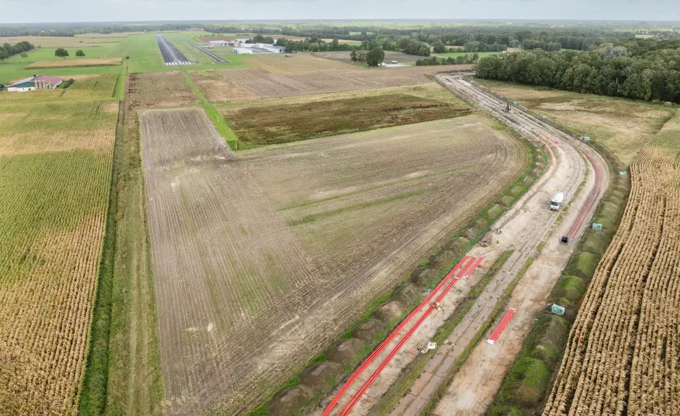 Luftaufnahme einer Großbaustelle für einen Erdkabelgraben, der durch landwirtschaftliche Felder verläuft, mit rotem Rohrmaterial und Baugeräten. Im Hintergrund sind die Landebahn und Gebäude eines kleinen Flugplatzes sichtbar, umgeben von Wald und Ackerland.