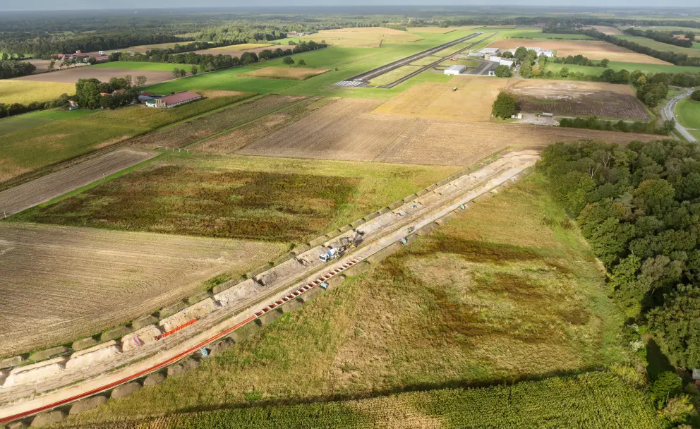 Luftaufnahme einer Baustelle für einen offenen Graben, der durch landwirtschaftliche Flächen und neben einem Flugplatz verläuft.