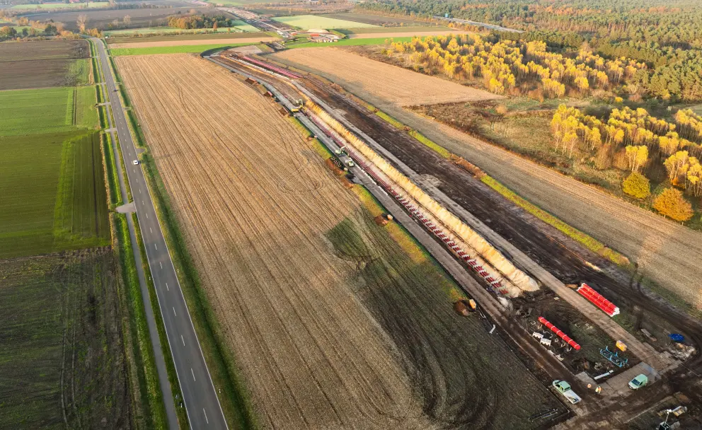 Herbstliche Drohnenaufnahme einer Kabelverlegetrasse: Auf einem abgeernteten Feld liegt eine lang gestreckte Baustelle mit aufgereihten Kabeltrommeln und Baufahrzeugen. Links verläuft eine Straße. Herbstlich verfärbte Bäume flankieren das rechte Ende der Trasse.