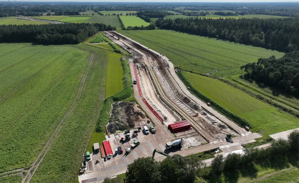 Sommertliche Drohnenaufnahme einer Kabelverlegebaustelle: Eine lang gestreckte Erdbaustelle mit Kabelgräben und roten Rohren verläuft durch eine Agrarlandschaft. Im Vordergrund ein Baustellenbereich mit Fahrzeugen und Containern, rechts ein Waldstreifen.