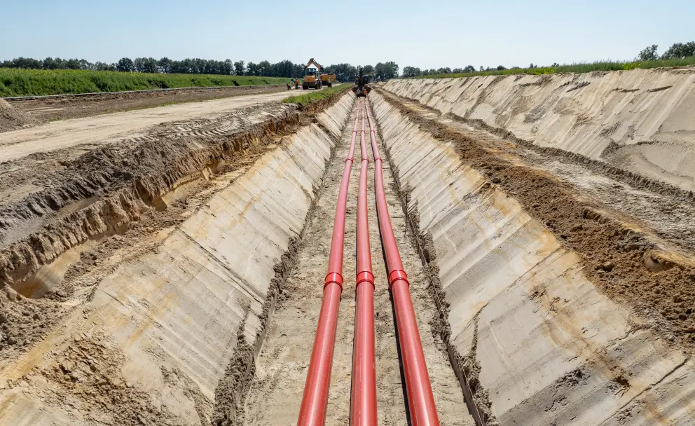 Bodennahe Aufnahme in einem geöffneten Kabelgraben:  parallel verlaufende rote Kabelschutzrohre liegen in einem tiefen, breiten Erdaushub. Im Hintergrund ist eine Baumaschine auf der Grabensohle zu sehen.