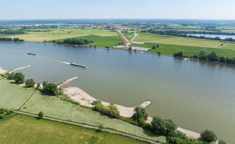 Drohnenaufnahme über dem Rhein: Zwei Frachtschiffe fahren auf dem breiten Fluss. Am gegenüberliegenden Ufer ist eine lang gestreckte Kabelverlegebaustelle erkennbar, die parallel zum Rhein verläuft. Im Hintergrund eine weite grüne Flusslandschaft mit Seen.