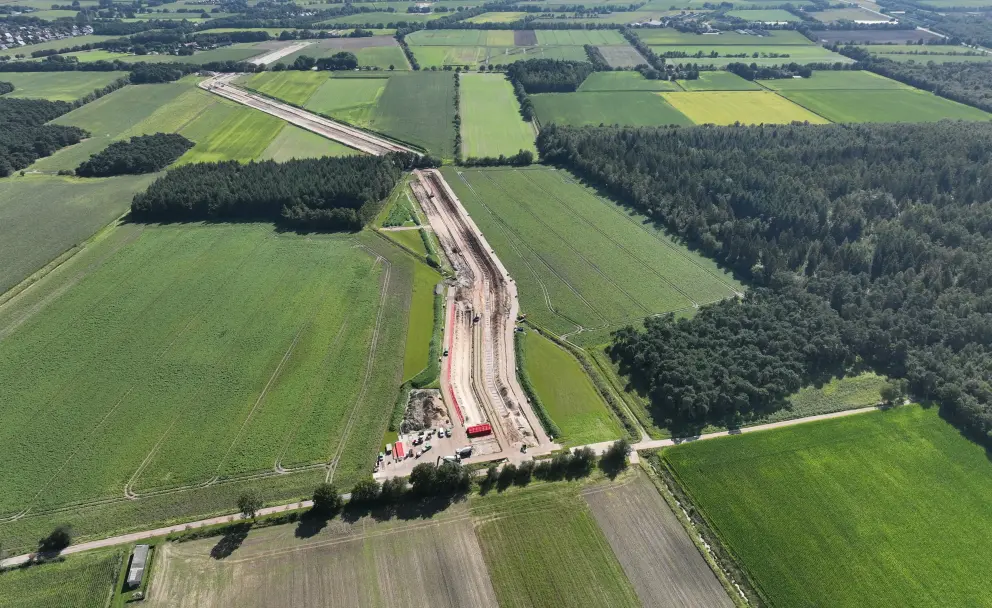 Drohnenaufnahme einer Kabelverlegebaustelle aus der Luft: Eine lang gestreckte Erdbaustelle mit roten Kabelschutzrohren verläuft durch eine norddeutsche Agrarlandschaft. Links grenzen Felder, rechts ein Waldstreifen an die Trasse.
