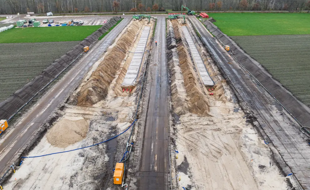 Drohnenaufnahme einer Kabelverlegebaustelle von oben: Ein breiter Erdeinschnitt mit zwei beidseits aufgeschütteten Erdwällen und einem befestigten Mittelstreifen erstreckt sich durch ein flaches Feld. Am Horizont ist ein Waldrand zu erkennen.