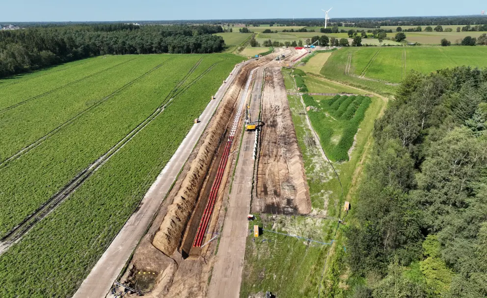 Drohnenaufnahme einer Kabelgrabungs-Baustelle im Sommer:  rote Kabelschutzrohre verlaufen in einem offenen Graben entlang einer Straße durch Felder. Rechts ist ein Waldstreifen zu sehen, im Hintergrund eine Windkraftanlage.