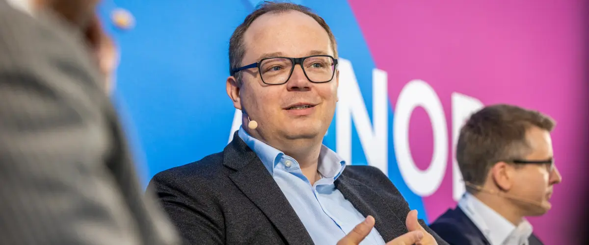 Dr. Jörn Koch spricht auf einem Podium mit lebhafter Gestik. Er trägt ein dunkles Sakko und ein Headset-Mikrofon. Im Hintergrund ist ein blau-magentafarbenes Veranstaltungsbanner zu sehen.
