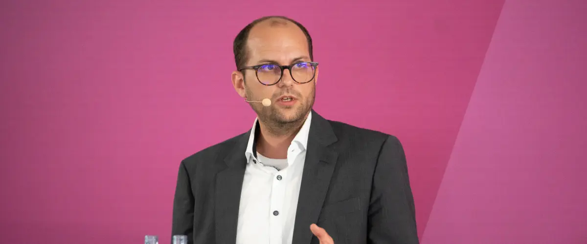 Markus Rützel spricht auf einem Podium vor einem magentafarbenen Hintergrund. Er trägt ein dunkles Sakko und ein Headset-Mikrofon. Links im Bild stehen Wasserflaschen.