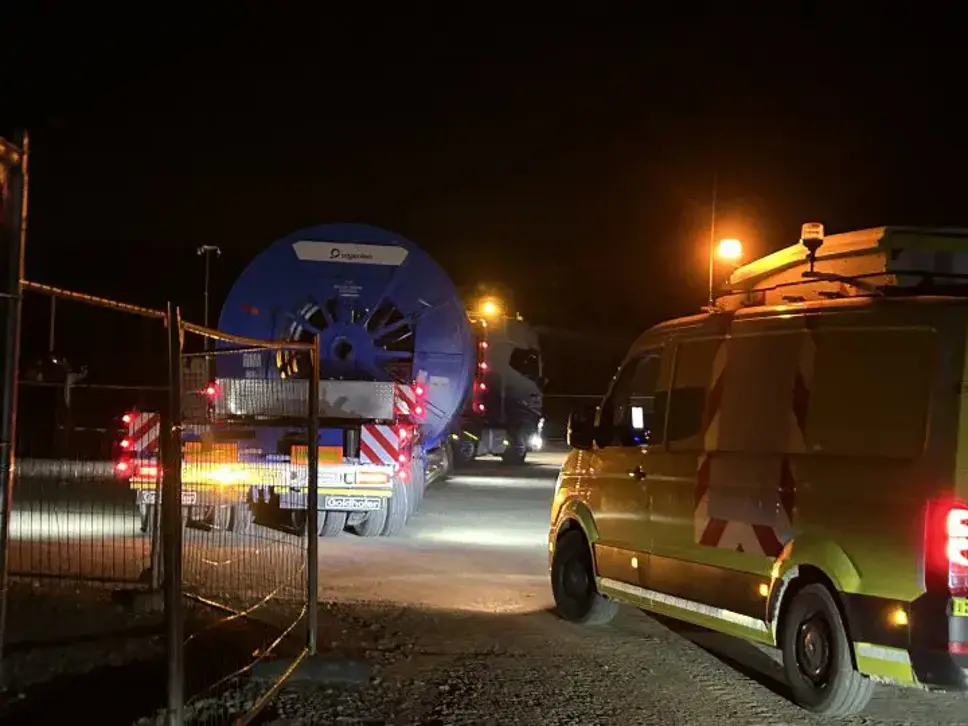 Nachtaufnahme: Ein Schwertransporter mit einer großen blauen Kabeltrommel (Amprion-Branding) fährt durch ein geöffnetes Baustellentor. Rechts daneben steht ein gelb-grünes Begleitfahrzeug.