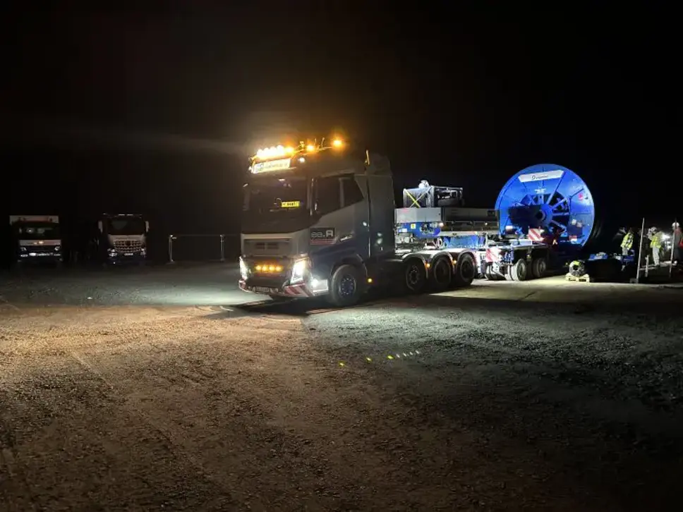 Nachtaufnahme auf einem Baustellengelände: Ein Schwertransporter steht beladen mit einer großen blauen Kabeltrommel (Amprion-Branding) auf einem unbefestigten Platz. Im Vordergrund ein weiterer Sattelzug mit eingeschalteten Scheinwerfern, rechts Monteure in Schutzkleidung.