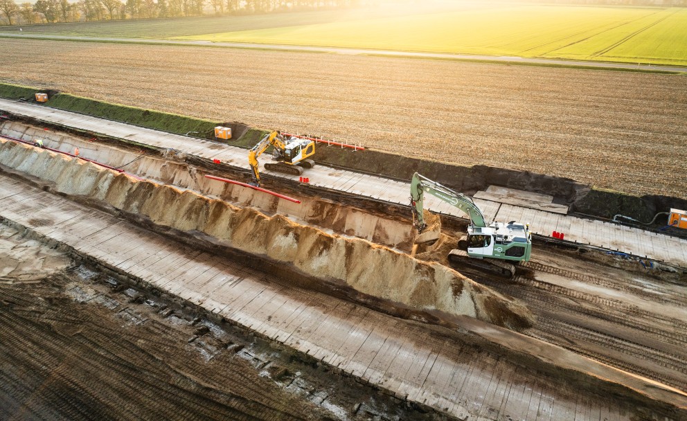 Luftaufnahme einer Baustelle mit zwei Baggern, die Erde in einem langen Graben bewegen, umgeben von Feldern im Sonnenlicht