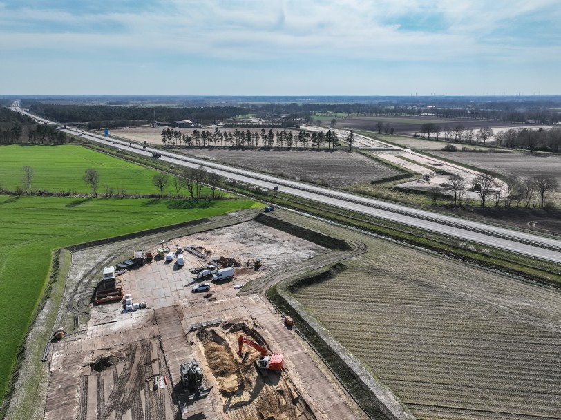 Luftaufnahme einer Baustelle an einer mehrspurigen Autobahn, die durch eine ländliche Region mit Feldern und Wäldern führt. Im Vordergrund steht ein Bagger an einer Erdaushubstelle.