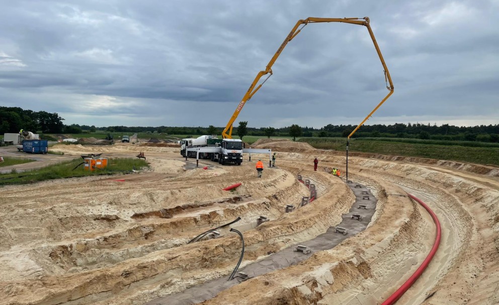 Großbaustelle mit einer mobilen Betonpumpe, deren langer, gelber Ausleger weit über eine halbkreisförmig terrassierte Baugrube ragt. Im Hintergrund steht ein bewölkter Himmel.