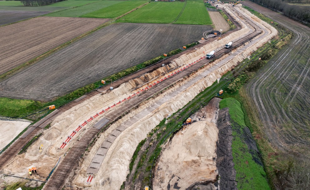 Luftaufnahme einer langgestreckten Baustelle, die sich kurvenförmig durch eine landwirtschaftliche Fläche zieht. In einem freigelegten Erdgraben ist eine lange Reihe roter Rohre verlegt.