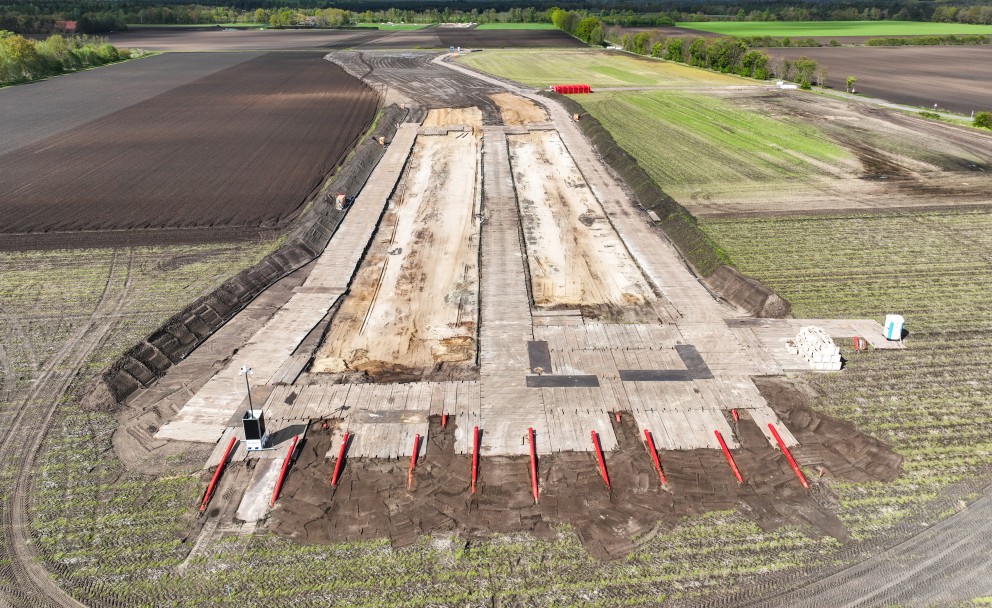 Luftaufnahme einer Baustelle auf einem Feld mit zwei großen, parallel verlaufenden, hellen Sandflächen. Im Vordergrund ragen mehrere rote Rohre schräg aus der Erde.