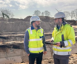 Zwei Personen mit Schutzhelmen und Warnwesten stehen auf einer Baustelle und unterhalten sich, eine hält ein Tablet