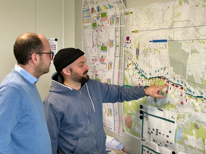 Zwei Männer stehen vor einer großen, detaillierten Karte an der Wand, einer zeigt mit dem Finger auf einen Bereich der Karte