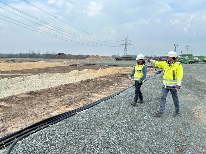 Zwei Bauarbeiter mit Schutzhelmen und Warnwesten gehen auf einer Baustelle mit Kabeln und Schotter, einer zeigt in die Ferne