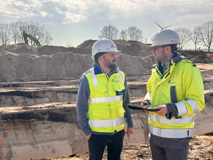 Zwei Personen mit Schutzhelmen und Warnwesten stehen auf einer Baustelle und unterhalten sich, eine hält ein Tablet