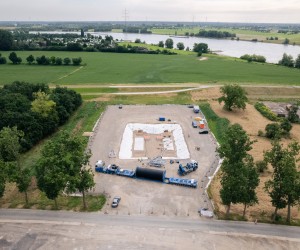 Luftaufnahme einer Baustelle mit rechteckigem Fundament neben einem Fluss und umgeben von Feldern und Bäumen
