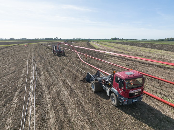 Inhalt: Luftaufnahme einer Großbaustelle auf einem flachen, braunen Feld unter blauem Himmel.Vordergrund: Ein dunkelroter MAN-Lkw mit Spezialreifen zieht eine Verlegevorrichtung durch die Erde.Hintergrund: Ein dickes, hellrotes Kabel wird in weiten Schleifen von einem hinteren Kettenfahrzeug über den Acker bis zum Lkw geführt. Links liegen bereits schmale, graue Leitungen parallel im Boden.Wenn Sie weitere Anpassungen für ein be