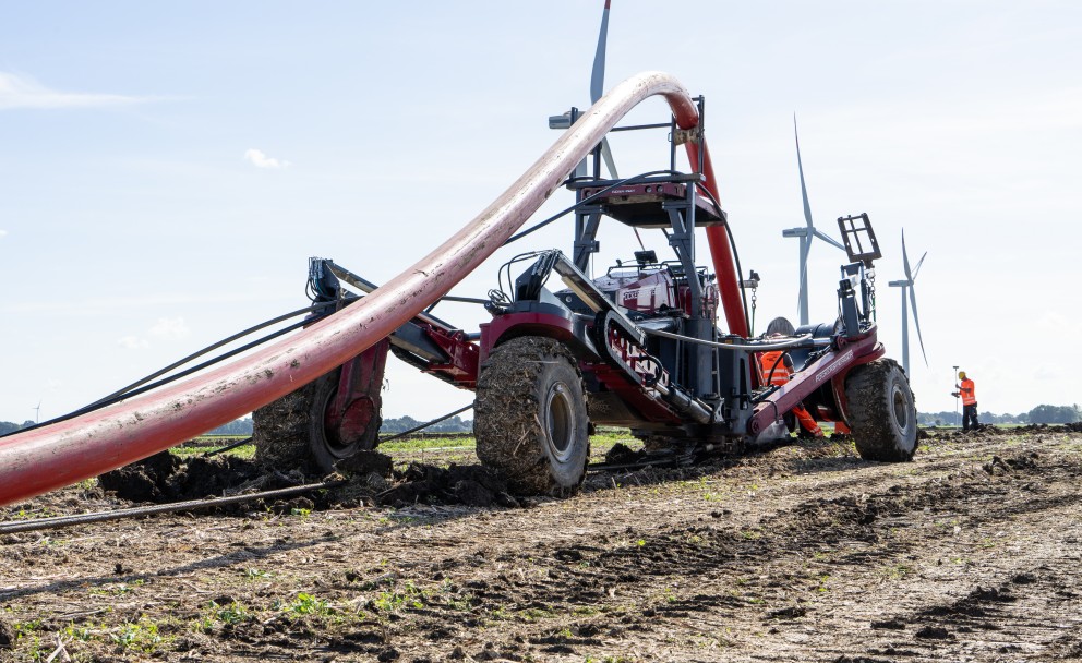 Vordergrund: Eine schwere Verlegemaschine mit großen, profilierten Reifen steht auf einem umgegrabenen Feld. Ein dickes, rotes Rohr oder Kabel wird von oben in die Führungsvorrichtung der Maschine geleitet.Hintergrund: Auf der flachen Landschaft im Hintergrund stehen mehrere Windkraftanlagen (Windräder). Ganz rechts steht eine Person in einer orangefarbenen Warnweste.