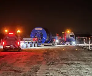 Nachtaufnahme auf einer verschneiten Baustelle: Ein Schwertransporter mit einer großen blauen Kabeltrommel steht auf einem schneebedeckten Untergrund. Im Vordergrund sind die roten Rücklichter eines weiteren Fahrzeugs sichtbar.