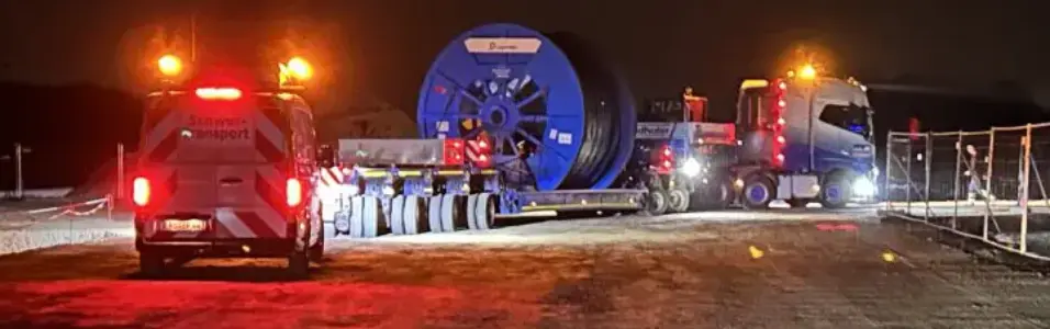 Nachtaufnahme auf einer verschneiten Baustelle: Ein Schwertransporter mit einer großen blauen Kabeltrommel steht auf einem schneebedeckten Untergrund. Im Vordergrund sind die roten Rücklichter eines weiteren Fahrzeugs sichtbar.