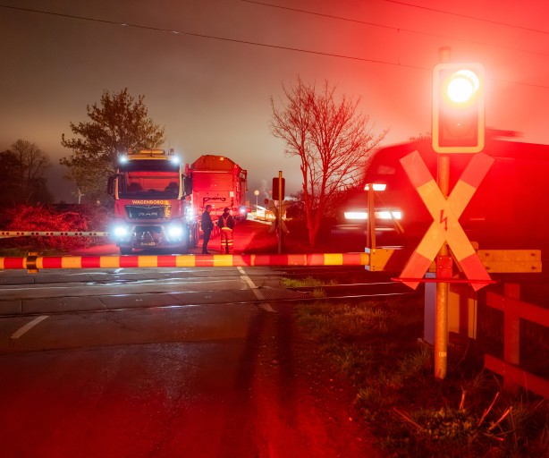 Geschlossener Bahnübergang bei Nacht mit rotem Warnlicht, einem wartenden Lastwagen und mehreren Personen auf der Straße
