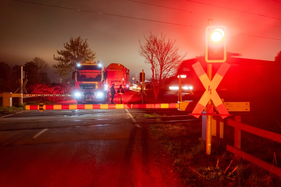 Geschlossener Bahnübergang bei Nacht mit rotem Warnlicht, einem wartenden Lastwagen und mehreren Personen auf der Straße