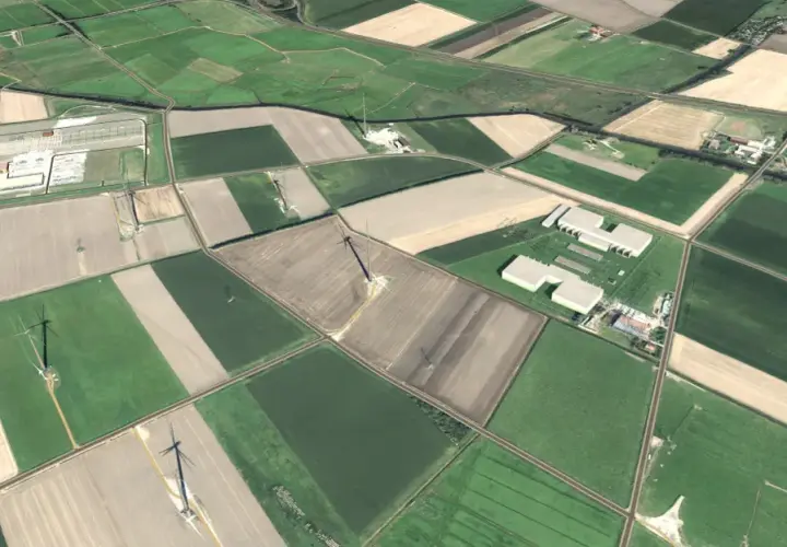 Luftaufnahme der Amprion Konverterstation Emden/Petkum während der Bauphase. Zu sehen sind mehrere landwirtschaftlich genutzte Flächen und die Betonstrukturen der im Bau befindlichen Konvertergebäude, umgeben von einem ländlichen Umfeld mit Wegen und vereinzelten Windkraftanlagen. 