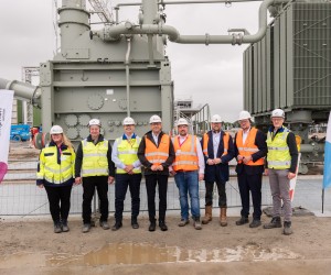 Eine Gruppe von neun Personen in Warnwesten und Schutzhelmen steht für ein Gruppenfoto nebeneinander auf einer Baustelle vor einer großen Industrieanlage. Links steht eine Werbefahne der Firma Amprion.
