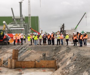 Eine große Gruppe von Personen in Warnwesten besichtigt eine Industrie-Großbaustelle. Im Hintergrund stehen eine massive graue Anlage, Kräne und ein roter Kettenbagger, während im Vordergrund ein ausgehobener Graben mit einer Treppe liegt.