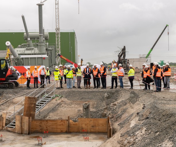 Eine große Gruppe von Personen in Warnwesten besichtigt eine Industrie-Großbaustelle. Im Hintergrund stehen eine massive graue Anlage, Kräne und ein roter Kettenbagger, während im Vordergrund ein ausgehobener Graben mit einer Treppe liegt.