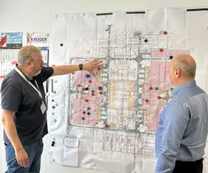 Zwei Männer in legerer Geschäftskleidung stehen vor einer großen technischen Zeichnung oder einem Bauplan, die an einer weißen Wand befestigt ist. Der Plan zeigt den Grundriss einer Anlage, wobei bestimmte Bereiche rosa schattiert und mit farbigen Magneten markiert sind.
Der Mann links trägt ein dunkelgraues Poloshirt, Bluejeans und ein Namensschild. Er deutet mit der Handfläche auf einen Bereich im linken Teil des Plans. Links neben dem Plan hängt ein Kalender und das Logo von "KRÜGER ELEKTROTECHNIK" und darunter "1150".
Der Mann rechts steht mit dem Rücken zur Kamera und trägt ein hellblaues Langarmhemd. Er betrachtet ebenfalls den Plan.
