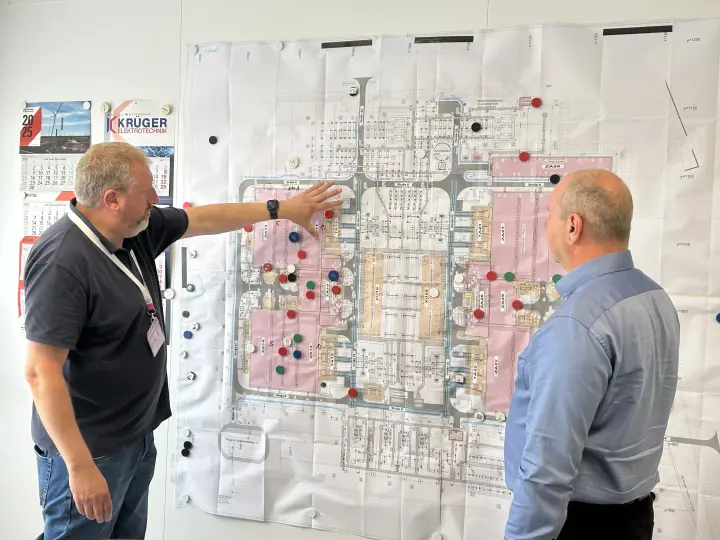 Zwei Männer in legerer Geschäftskleidung stehen vor einer großen technischen Zeichnung oder einem Bauplan, die an einer weißen Wand befestigt ist. Der Plan zeigt den Grundriss einer Anlage, wobei bestimmte Bereiche rosa schattiert und mit farbigen Magneten markiert sind.
Der Mann links trägt ein dunkelgraues Poloshirt, Bluejeans und ein Namensschild. Er deutet mit der Handfläche auf einen Bereich im linken Teil des Plans. Links neben dem Plan hängt ein Kalender und das Logo von "KRÜGER ELEKTROTECHNIK" und darunter "1150".
Der Mann rechts steht mit dem Rücken zur Kamera und trägt ein hellblaues Langarmhemd. Er betrachtet ebenfalls den Plan.
