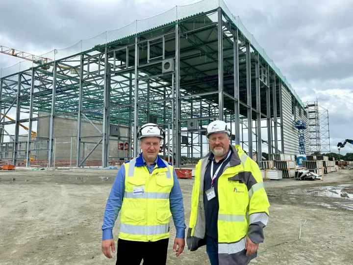 Zwei Männer in Warnwesten, Schutzhelmen und blauen Hemden posieren auf der Baustelle der Amprion Konverterstation A-Nord in Emden-Petkum.
Der linke Mann trägt eine gelbe Warnweste, der rechte eine gelbe Warnjacke mit grauen Details und einem Amprion-Logo. Im Hintergrund befindet sich die im Bau befindliche, weitläufige Stahlkonstruktion der Konverterhalle, umgeben von Baumaschinen und Material. Diese Station ist ein wesentlicher Bestandteil der Energiewende, da sie Windstrom aus der Nordsee für den Transport per Erdkabel ins Gleichstromnetz umwandeln wird.