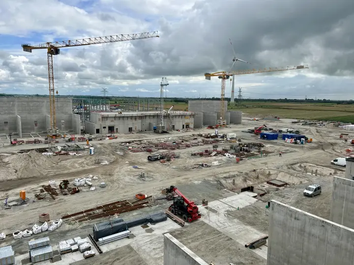 Das Bild zeigt eine weitläufige Industriebaustelle unter einem bewölkten Himmel. Im Fokus stehen mehrere im Bau befindliche Betonstrukturen, die die zukünftigen Gebäude der Konverterstation bilden.
Krane und Ausrüstung: Auf dem Gelände sind zwei große, gelbe Turmdrehkrane positioniert. Ein dritter, kleinerer Kran ist in der Mitte sichtbar. Am Boden sind diverse Baumaschinen, darunter ein roter Raupenbagger, und Baumaterialien wie Betonfertigteile und Bewehrungsstahl verteilt.
Hintergrund und Umgebung: Im Hintergrund, hinter der Baustelle und auf angrenzenden Feldern, sind Freileitungsmasten und Windkraftanlagen zu erkennen, was die Verbindung des Projekts zur Energieversorgung unterstreicht. Das umliegende Gebiet besteht aus flachem, ländlichem Ackerland.
Diese Anlage ist ein zentraler Bestandteil der Energiewende und wird dazu dienen, in der Nordsee erzeugten Windstrom über die Erdkabeltrasse A-Nord in das deutsche Stromnetz einzuspeisen.