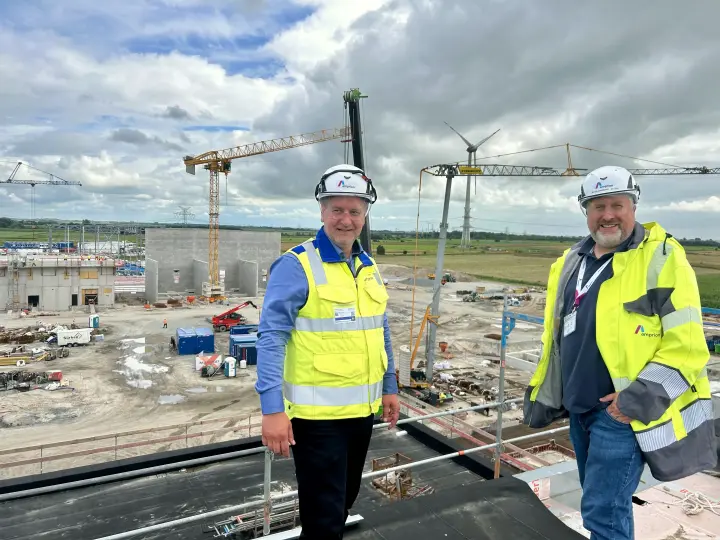 Zwei Männer in Schutzkleidung stehen auf dem Dach einer großen Baustelle mit Kränen und Windrädern im Hintergrund.