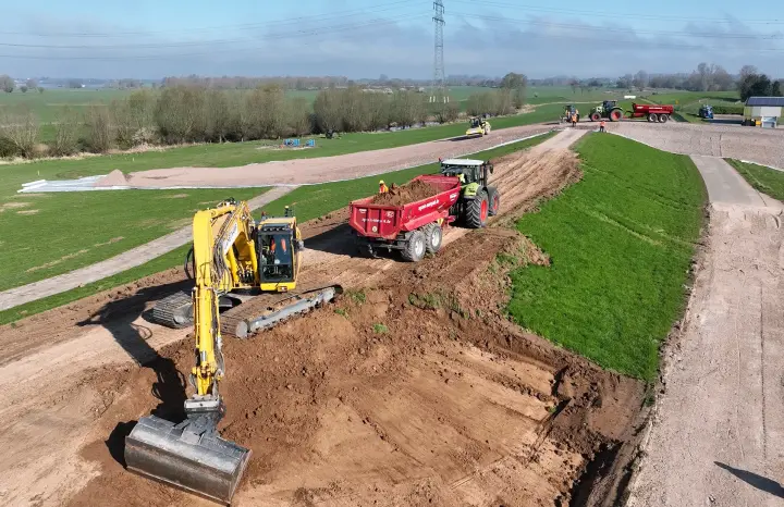 Das Bild zeigt eine aktive Baustelle, auf der Erdarbeiten durchgeführt werden.
Vordergrund: Im Vordergrund ist ein großer, gelber Raupenbagger zu sehen, der Erdreich bewegt und einen tiefen, offenen Graben aushebt. Rechts neben dem Bagger steht ein roter Muldenkipper-Anhänger (Halfpipe-Mulde), der von einem großen grünen Traktor gezogen wird.
Mittelgrund: Eine erhöhte Erdstraße führt durch die Landschaft. Weiter hinten, auf diesem provisorischen Weg, sind weitere Baumaschinen aktiv: eine gelbe Walze und ein weiterer Traktor mit einem Anhänger. Personen in Warnkleidung sind in der Nähe der Fahrzeuge zu erkennen.
Hintergrund: Die Baustelle verläuft durch grüne Wiesenflächen und ist von dichter Busch- und Baumvegetation begrenzt. Im oberen Teil des Bildes sind markante Freileitungsmasten und Stromleitungen sichtbar. 