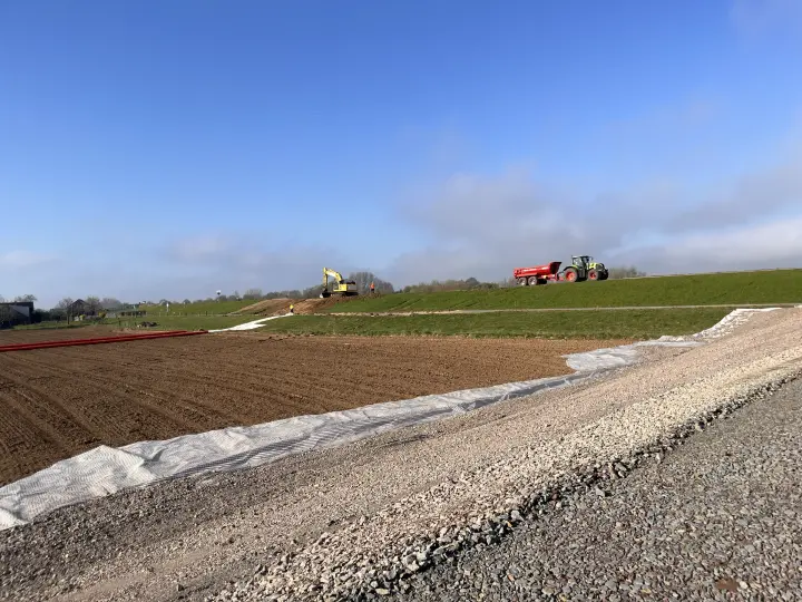 Eine Baustelle für ein großes Infrastrukturprojekt, vermutlich eine Erdkabeltrasse, erstreckt sich durch eine ländliche, von Deichen geprägte Landschaft. Das Bild wurde aus einer niedrigen Perspektive aufgenommen, die den Blick auf einen massiven, frisch angelegten Schotterwall im Vordergrund lenkt. Dieser Wall ist teilweise mit einem weißen Geotextil bedeckt, um Erosion zu verhindern.
Baustellenaktivität und Umgebung
Vorder- und Mittelgrund: Dahinter liegt ein großes, braunes, umgepflügtes Feld. Auf der linken Seite dieses Feldes sind lange, rote Schutzrohre sichtbar, die für die Verlegung der Kabel vorbereitet wurden.
Hintergrund: Auf einem erhöhten grünen Deich oder Damm im mittleren Bereich des Bildes sind zwei schwere Baumaschinen im Einsatz:
Links arbeitet ein gelber Kettenbagger.
Rechts fährt ein großer grüner Traktor mit einem roten Kippanhänger (Muldenkipper), beladen mit Material.
Atmosphäre: Der Himmel ist strahlend blau mit nur wenigen, weit entfernten Wolken, was auf gutes Bauwetter hinweist. Die Szene vermittelt den Eindruck eines großflächigen, aktiven Bauvorhabens in einer landwirtschaftlich genutzten Region.