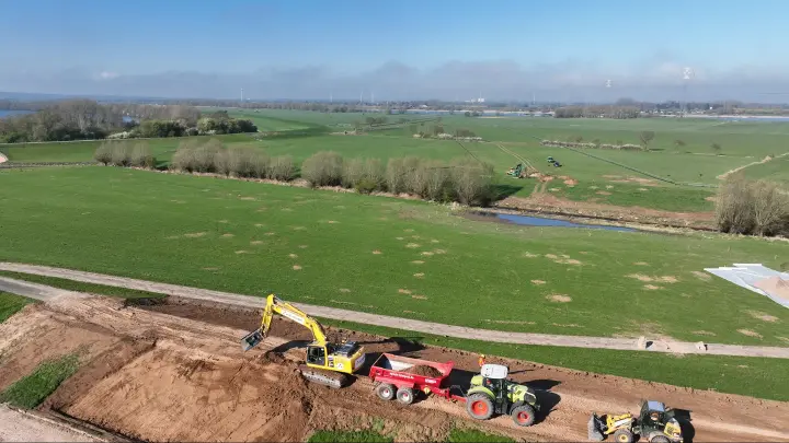 Dies ist eine Luftaufnahme einer Großbaustelle, auf der ein gelber Raupenbagger und ein grüner Traktor mit einem roten Muldenkipper im Einsatz sind. Die Szene ist typisch für Bauarbeiten im Rahmen großer Infrastrukturprojekte, wie der Verlegung von Erdkabeln für die Stromautobahn A-Nord durch das ländliche Deutschland.